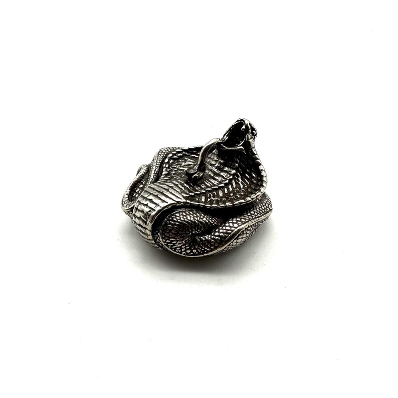 May include: A silver-coloured cobra figurine, coiled and ready to strike. The snake's scales are intricately textured, and its hood is raised. The figurine is set against a plain white background, highlighting the detailed craftsmanship.