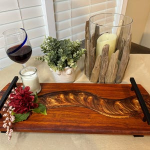 May include: A wooden serving tray with a gold and brown epoxy river design. The tray has black metal handles and is decorated with a red flower and greenery.