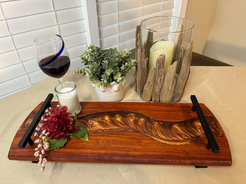 May include: A wooden serving tray with a gold and brown epoxy river design. The tray has black metal handles and is decorated with a red flower and greenery.