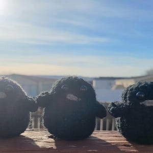 May include: Three black knitted penguin figurines with white beaks and black eyes. The penguins are sitting on a wooden surface.