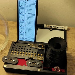 Könnte beinhalten: Ein schwarzes und goldenes Tischspielset für Dungeons and Dragons Rollenspiele. Das Set enthält einen Telefonständer, einen Würfelturm, einen Zauberplatz-Tracker, einen Konzentrations-Tracker und einen Todesrettungs-Tracker. Der Telefonständer hat einen Bildschirm, der ein Charakterblatt mit Statistiken und Rettungswürfen anzeigt. Der Würfelturm ist aus schwarzem Kunststoff und hat eine zylindrische Form. Der Zauberplatz-Tracker hat ein Raster mit neun Knöpfen mit den Zahlen 1 bis 9. Der Konzentrations-Tracker hat zwei Knöpfe mit den Beschriftungen "Ja" und "Nein". Der Todesrettungs-Tracker hat drei Knöpfe mit den Beschriftungen "1", "2" und "+".