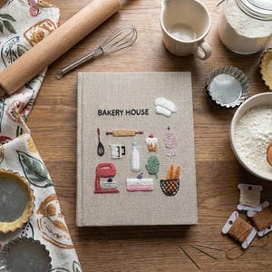 May include: A cookbook titled "Bakery House" with embroidered illustrations of baking tools and treats. The image includes a rolling pin, whisk, measuring cup, flour, eggs, and pie tins, all arranged on a wooden surface, suggesting a baking theme.