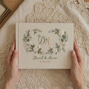 May include: A cream-colored wedding guest book with embroidered floral wreath and initials "D & A". The book cover also features the names "David & Anna" and the date "13.09.2025" in gold and green thread.