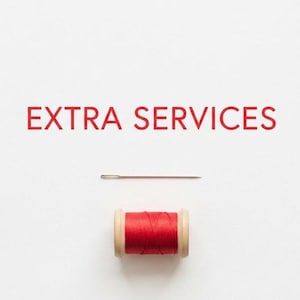 May include: The text EXTRA SERVICES is printed in red capital letters. Below the text is a silver sewing needle positioned horizontally. Beneath the needle is a wooden spool wrapped with red sewing thread on a plain white background.