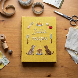 May include: A yellow fabric-covered book with the embroidered text "Julia recipes" in white. The cover features embroidered illustrations of a whisk, a plate of food, a tomato, a fork, a knife, a stack of pancakes, and two brown dogs.
