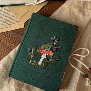 以下が含まれることがあります： 赤と白のキノコにカエルが刺繍された、ベリーと葉で囲まれた濃い緑色のジャーナル。ジャーナルには装飾的なコーナープロテクターとクリーム色の紐留めが付いています。