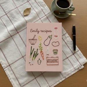 May include: A pink recipe book titled "Emily recipes" with embroidered illustrations of vegetables and kitchen items. A cup of coffee, a pen, and a checkered cloth are also in the image.