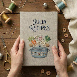May include: A light beige fabric-covered book with the embroidered text JULIA RECIPES. The cover features an embroidered blue pot with a floral pattern, surrounded by stitched vegetables including carrots, mushrooms, an onion, and a leek.