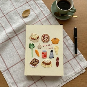 Puede incluir: Un cuaderno beige con ilustraciones bordadas de comida, incluyendo una pizza, una olla y verduras. El texto "DINNER TIME" está bordado en la portada. Una taza de café, un bolígrafo y una cuchara están sobre la mesa.
