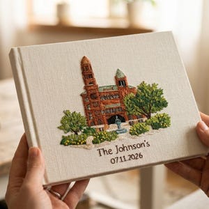 Puede incluir: Un libro rectangular cubierto de tela color crema con un diseño bordado de un edificio de ladrillo con una torre, árboles verdes y una fuente. Debajo del bordado, el texto dice The Johnson's y la fecha 07.11.2026.