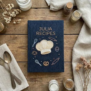 May include: A navy blue recipe book with the title "JULIA RECIPES" embroidered in gold thread. The cover features a chef's hat and cooking-themed illustrations. The book is surrounded by cooking utensils and spices on a wooden surface.