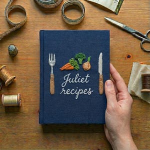 May include: A navy blue fabric-covered journal with embroidered vegetables, a fork, and a knife on the front cover. The text "Juliet recipes" is stitched in white thread in the center. The book is surrounded by sewing supplies on a wooden surface.