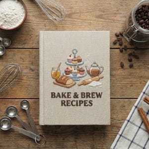 May include: A beige cookbook titled "BAKE & BREW RECIPES" with an embroidered illustration of cupcakes, bread, and a teapot. Baking and brewing tools, including measuring spoons, a whisk, and a jar of coffee beans, surround the book on a wooden surface.