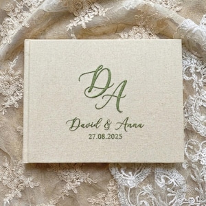 Può includere: Un libro degli ospiti rettangolare rivestito in tessuto beige chiaro con ricamo verde oliva. La copertina presenta il monogramma DA, i nomi David & Anna e la data 27.08.2025 in un carattere corsivo centrato sulla parte anteriore.