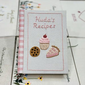 May include: Embroidered recipe book titled "Huda's Recipes" with a cupcake, cookie, and pie slice design. The book has a light pink cover with a gingham spine. The cover features embroidered images of baked goods.