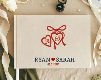 May include: A cream-colored wedding guest book with red embroidered hearts and a bow design. The names "Ryan" and "Sarah" are printed in navy blue, with a red heart symbol between them, and the date "03.01.2025" below. A white ribbon bookmark is visible.