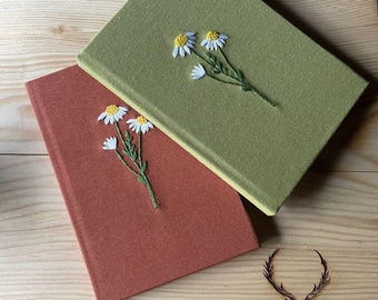 May include: Two hardback books, one olive green and one rust-coloured, are shown. Each book has an embroidered daisy floral design. The books are set on a wooden surface, with a deer head logo and the word "KEBIKEÇ" etched into the wood.