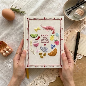 May include: A white journal with embroidered food illustrations, including a shrimp, lemon, and croissant. The cover features the text "Enjoy the Little Things" in red. The journal is held by hands.