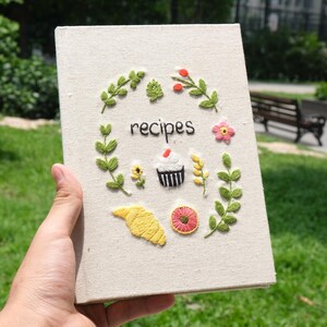 May include: A white linen recipe book with embroidered flowers, leaves, a cupcake, and the word "recipes".