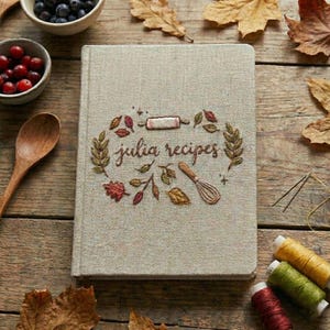 May include: A linen-covered recipe book with embroidered details. The cover features the text "julia recipes" surrounded by a wreath of leaves, a rolling pin, and a whisk. The book is on a wooden surface with fall foliage and bowls of berries.
