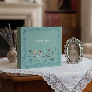 May include: A teal-colored box with the text "NOVO ROSE & RAFAEL" and embroidered embellishments including a rainbow, butterflies, and musical notes. The box is on a table with a lace doily, next to a framed photograph.