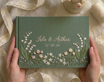 Könnte beinhalten: Ein salbeigrünes, stoffbezogenes Buch mit gestickten weißen Maiglöckchen und kleinen rosa Blumen. In der Mitte befindet sich der gestickte Text Isla & Arthur sowie das Datum 27.07.24 in einer weißen Schreib- und Blockschrift.
