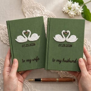 Puede incluir: Dos libros de tapa dura color verde oliva con motivos de cisnes bordados en blanco, la fecha 25.07.2026 y el texto to my wife y to my husband. Una pluma estilográfica de madera marrón está colocada horizontalmente bajo los libros.