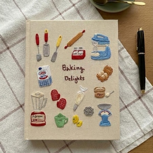 Puede incluir: Una cubierta de diario beige bordada con varias imágenes temáticas de repostería, incluyendo una batidora, un rodillo y huevos. El texto "Baking Delights" está bordado en la cubierta. Un bolígrafo negro descansa sobre un paño a cuadros blanco y marrón.