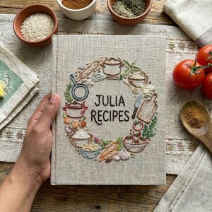 May include: A linen-covered cookbook titled "Julia Recipes" with an embroidered wreath of cooking tools and ingredients. The book is surrounded by small bowls of spices, tomatoes, and a wooden spoon, suggesting a culinary theme.