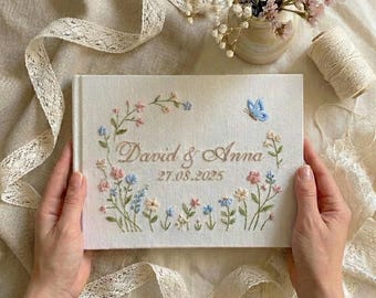 May include: A rectangular white fabric-covered book featuring embroidered floral motifs in shades of pink and blue, a small blue butterfly, and the embroidered text David & Anna 27.08.2025 centered on the front cover with a clean finish.