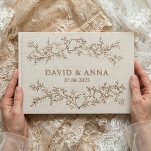 Può includere: Un album di nozze beige in lino con ricami floreali. La copertina presenta i nomi "DAVID & ANNA" e la data "27.08.2025" in marrone. L'album è posto su uno sfondo di tessuto di pizzo.