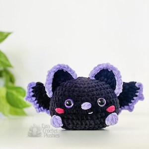 Puede incluir: Un murci&eacute;lago de peluche tejido a crochet con alas moradas y un cuerpo negro. El murci&eacute;lago tiene mejillas rosadas y ojos negros. El murci&eacute;lago est&aacute; sentado sobre una superficie blanca.