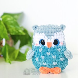 Puede incluir: Un juguete de peluche de b&uacute;ho de crochet azul con detalles blancos alrededor de los ojos y un pico naranja brillante. El b&uacute;ho tiene grandes ojos azules con pupilas negras. El juguete est&aacute; sentado sobre una superficie blanca.