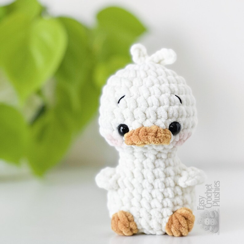 Peut inclure: Un petit canard en peluche crochet&eacute; blanc avec une finition en laine textur&eacute;e. Il pr&eacute;sente un bec beige, deux yeux de s&eacute;curit&eacute; noirs, de petites ailes et des pieds beiges. La t&ecirc;te a une petite touffe de laine, et le texte Easy Crochet Plushies est visible.