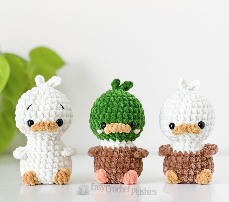 May include: Three handmade crochet duck plushies. One is white, one is green, and one is white. Each has a brown beak and feet, and black eyes. The green duck has a green head and brown body. The white ducks have white heads and brown bodies.