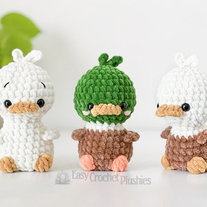 Peut inclure: Trois petites peluches crochet&eacute;es &agrave; la main en forme de canards. L'une est enti&egrave;rement blanche, une a une t&ecirc;te verte avec un corps marron, et une a une t&ecirc;te blanche avec un corps marron. Chacune poss&egrave;de des yeux noirs, un bec beige et des joues roses.