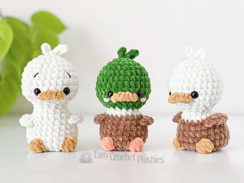 Peut inclure: Trois petites peluches crochet&eacute;es &agrave; la main en forme de canards. L'une est enti&egrave;rement blanche, une a une t&ecirc;te verte avec un corps marron, et une a une t&ecirc;te blanche avec un corps marron. Chacune poss&egrave;de des yeux noirs, un bec beige et des joues roses.