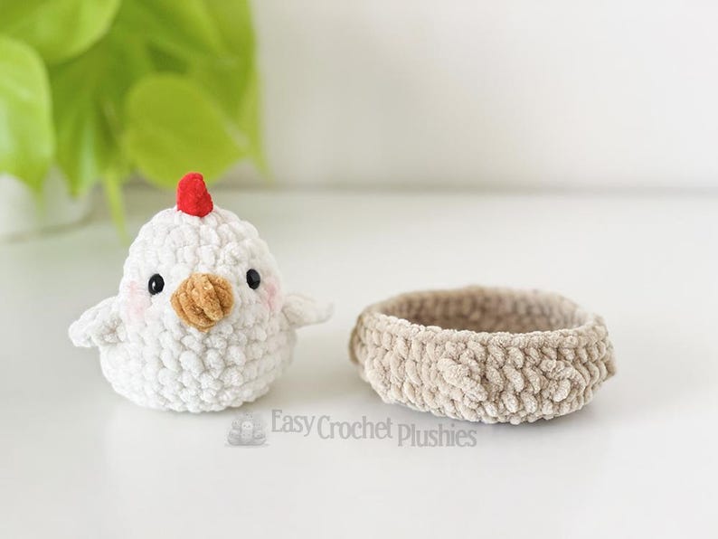 May include: A white crochet chick with a red comb and a tan beak sits beside a small, tan crochet basket. The chick has black eyes and pink cheeks. The items are handmade and suitable for Easter.