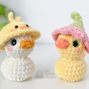 May include: Two handmade crochet plushies of ducklings. One is white with a yellow hat, the other is yellow with a pink and green hat. Both have orange beaks, rosy cheeks, and sparkly eyes. The text "Easy Crochet Plushies" is visible.