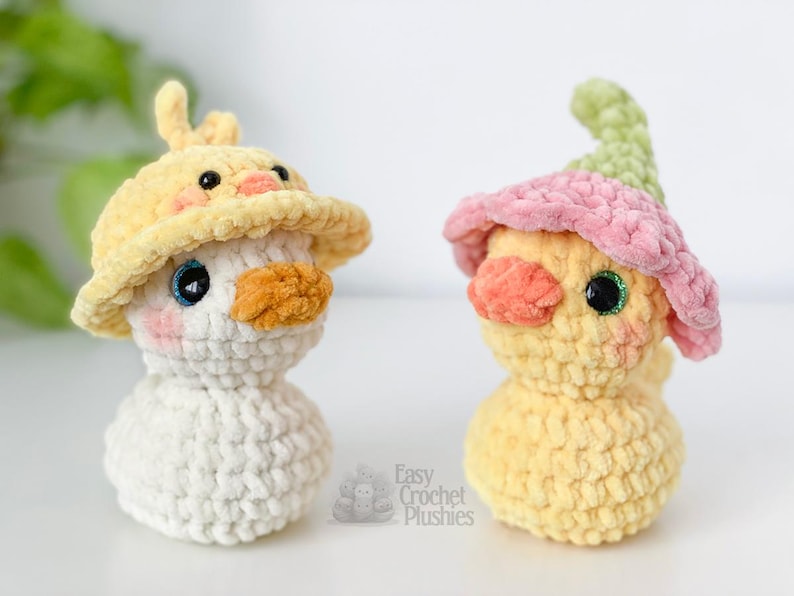 May include: Two handmade crochet plushies of ducklings. One is white with a yellow hat, the other is yellow with a pink and green hat. Both have orange beaks, rosy cheeks, and sparkly eyes. The text "Easy Crochet Plushies" is visible.
