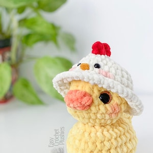 May include: A handmade yellow crochet chick plushie wearing a white hat with a red comb. The chick has a peach-colored beak and rosy cheeks. The plushie is a soft, textured design, perfect for a child's toy or nursery decor.