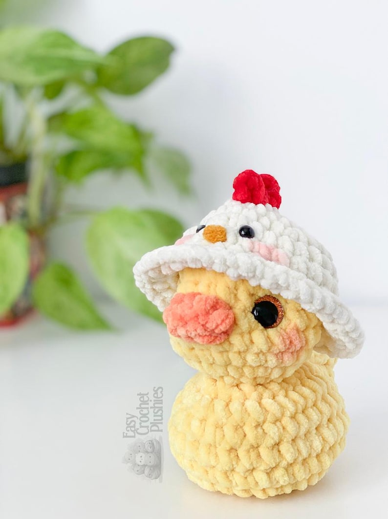 May include: A handmade yellow crochet chick plushie wearing a white hat with a red comb. The chick has a peach-colored beak and rosy cheeks. The plushie is a soft, textured design, perfect for a child's toy or nursery decor.