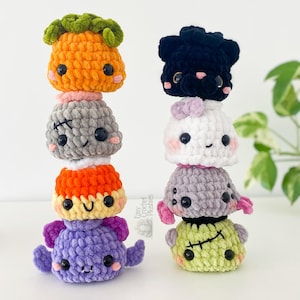 May include: Two vertical stacks of eight small, round, crocheted plushies. The left stack features an orange pumpkin, a gray monster with stitches, a yellow character, and a purple bat. The right stack includes a black cat, a white ghost, a gray spider, and a green monster with stitches. Each plush has black plastic eyes and pink cheeks.