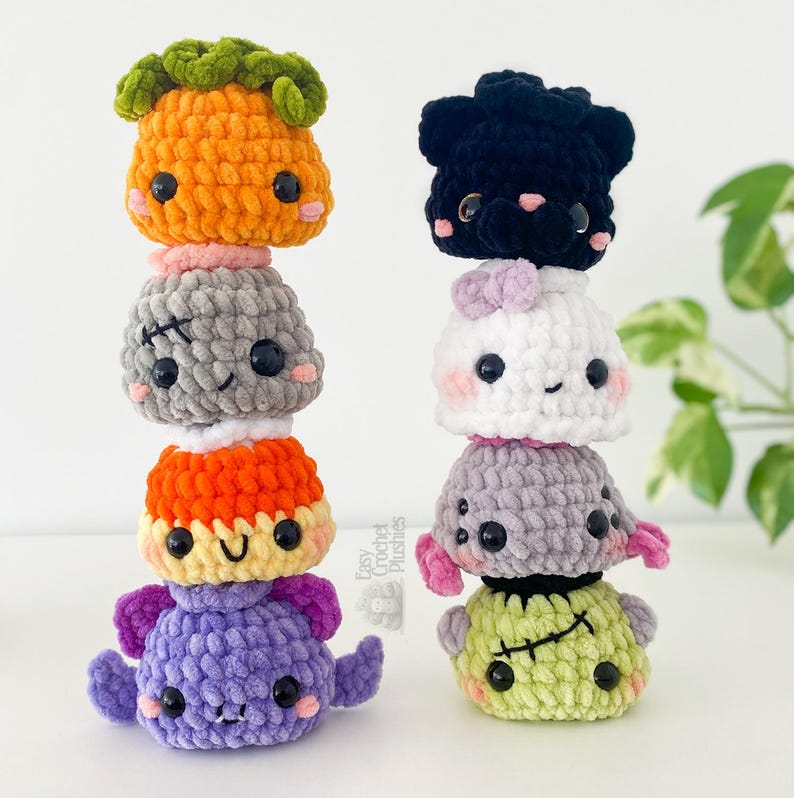 May include: Two vertical stacks of eight small, round, crocheted plushies. The left stack features an orange pumpkin, a gray monster with stitches, a yellow character, and a purple bat. The right stack includes a black cat, a white ghost, a gray spider, and a green monster with stitches. Each plush has black plastic eyes and pink cheeks.