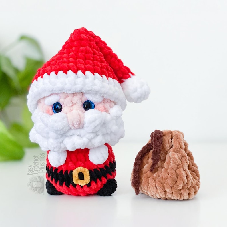 May include: A handmade crochet Santa Claus plushie with a red hat, white beard, and black belt. A small brown drawstring bag sits beside the Santa figure. The plushie has blue eyes and is made of soft yarn. The text "Easy Crochet Plushies" is visible.