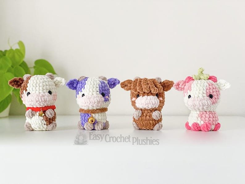 May include: Four handmade crochet cow plushies in various colours: brown and white, purple and white, brown, and pink and white. Each plush has a unique design, including bells and a strawberry-like top. The text "Easy Crochet Plushies" is visible.