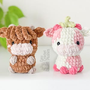 May include: Two crochet cow plushies. One is brown with a white muzzle and ears. The other is pink and white with a green stem on its head. Both have black eyes and small arms and legs.