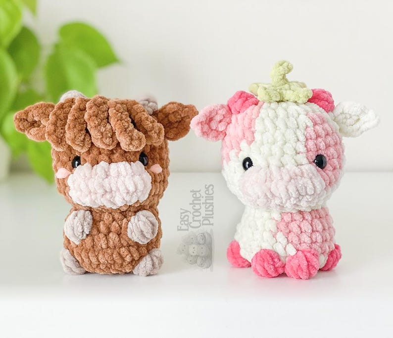 May include: Two crochet cow plushies. One is brown with a white muzzle and ears. The other is pink and white with a green stem on its head. Both have black eyes and small arms and legs.