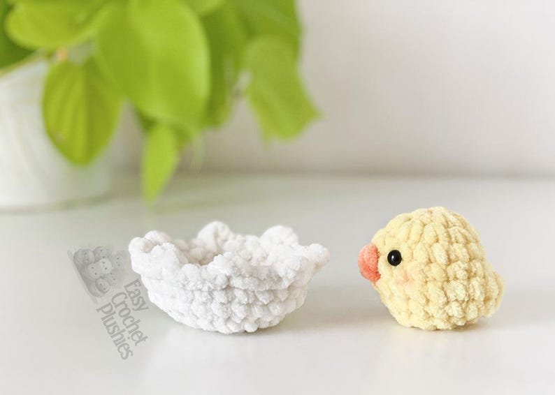 May include: A crocheted yellow chick with a peach beak and black eyes sits beside a white crocheted bowl. The chick has rosy cheeks. The bowl is round with a ruffled edge. The text "Easy Crochet Plushies" is visible.