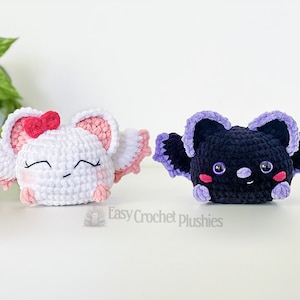 Puede incluir: Dos juguetes de peluche de crochet, uno blanco con detalles rosas y un lazo rosa, y otro negro con detalles morados. Ambos juguetes tienen ojos bordados y una boca sonriente.